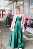 A person in a flowing emerald green dress, complemented by a stylish pink blazer. The individual is standing in an urban environment, holding a small white handbag. The background includes elements such as a trash bin, shopping carts, and a storefront.