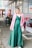 A person in a flowing emerald green dress, complemented by a stylish pink blazer. The individual is standing in an urban environment, holding a small white handbag. The background includes elements such as a trash bin, shopping carts, and a storefront.