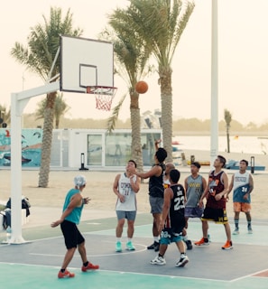 A group of people are playing basketball on an outdoor court near a beach setting. Palm trees are in the background, and there is a mural on a nearby wall. Some players are in mid-action, with one attempting a shot towards the hoop.
