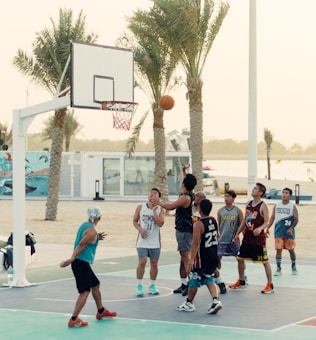 A group of people are playing basketball on an outdoor court near a beach setting. Palm trees are in the background, and there is a mural on a nearby wall. Some players are in mid-action, with one attempting a shot towards the hoop.