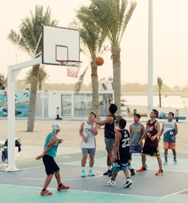 A group of people are playing basketball on an outdoor court near a beach setting. Palm trees are in the background, and there is a mural on a nearby wall. Some players are in mid-action, with one attempting a shot towards the hoop.