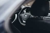 A close-up view of a vehicle's interior focusing on the steering wheel, which features a BMW logo. The dashboard and part of the driver's seat are also visible, with a sleek and modern design.