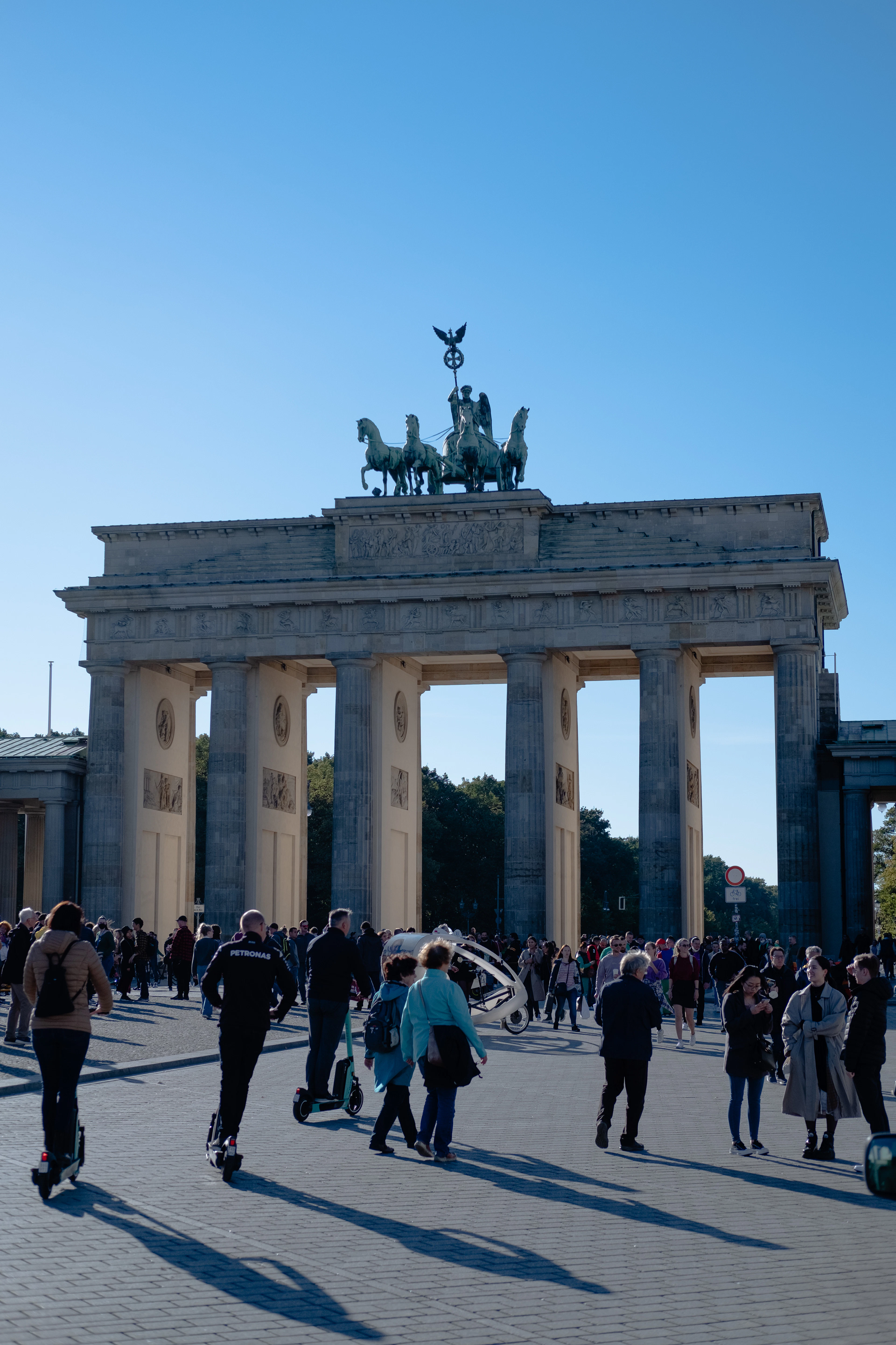 The Brandenburg Gate in Berlin on a clear day, its Quadriga chariot statue on top and crowds of people crossing Pariser Platz in front of the columns.