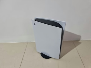 A sleek, modern gaming console stands vertically on a light-colored tiled floor. Featuring a distinctive white and black design, the console has a curved, futuristic appearance with the PlayStation logo visible on the front panel.