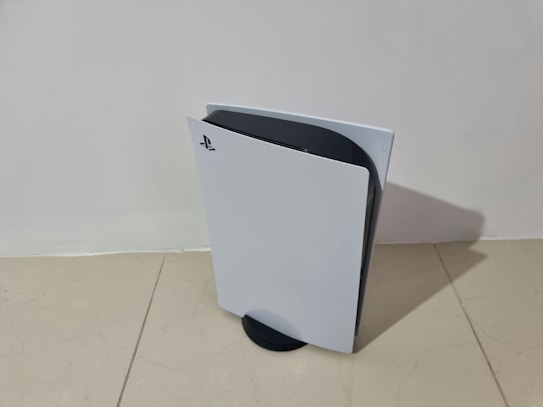 A sleek, modern gaming console stands vertically on a light-colored tiled floor. Featuring a distinctive white and black design, the console has a curved, futuristic appearance with the PlayStation logo visible on the front panel.