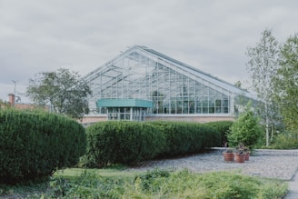 A large greenhouse with a glass exterior is surrounded by neatly trimmed hedges and various green plants. The structure has a modern design with a sloped roof and is situated in a landscaped garden area that includes small trees and decorative pots. The sky above is cloudy, creating a calm atmosphere.