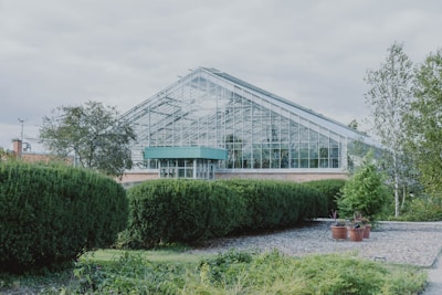 A large greenhouse with a glass exterior is surrounded by neatly trimmed hedges and various green plants. The structure has a modern design with a sloped roof and is situated in a landscaped garden area that includes small trees and decorative pots. The sky above is cloudy, creating a calm atmosphere.