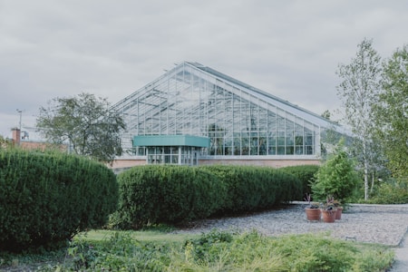 A large greenhouse with a glass exterior is surrounded by neatly trimmed hedges and various green plants. The structure has a modern design with a sloped roof and is situated in a landscaped garden area that includes small trees and decorative pots. The sky above is cloudy, creating a calm atmosphere.