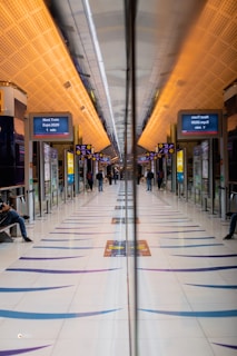 A modern train station platform with a symmetrical design featuring a reflective surface in the middle, leading to a vanishing point. The ceiling has a warm, orange lighting, and digital signs display train information. Several people are visible, some sitting and some walking.