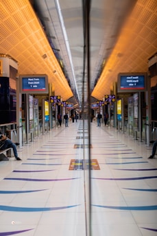 A modern train station platform with a symmetrical design featuring a reflective surface in the middle, leading to a vanishing point. The ceiling has a warm, orange lighting, and digital signs display train information. Several people are visible, some sitting and some walking.