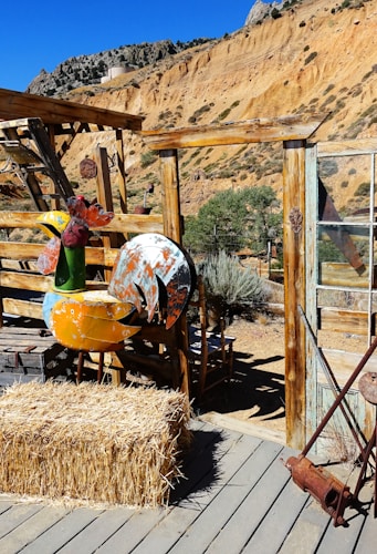 A rustic outdoor setting features a wooden structure with a hay bale placed on a wooden deck. In the background, rugged orange-brown cliffs rise against a bright blue sky, and a large metal sculpture resembling a bird stands next to the hay bale.