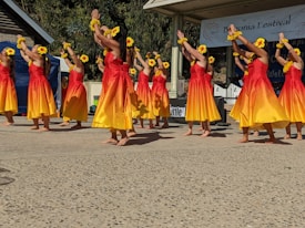 A group of dancers wearing yellow and red dresses with yellow flowers in their hair performs on an outdoor stage. Their arms are raised, and they are moving in unison. The background shows a banner for a festival and some trees.