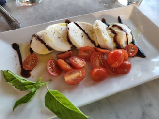 Slices of fresh mozzarella cheese are drizzled with balsamic glaze and placed alongside cherry tomatoes on a rectangular white plate. Fresh basil leaves are arranged on the side, adding a touch of green to the dish.