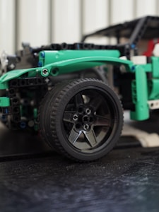 Close-up view of a detailed model car made from interlocking plastic building blocks. The focus is on a black tire with a five-spoke rim, surrounded by green and black components of the model.