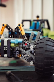 A close-up of a model car assembled from interlocking plastic bricks. The focus is on the front suspension and large tire, featuring intricate details like yellow struts and visible connectors. The background shows additional components and parts of the car, slightly out of focus.