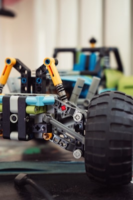 A close-up of a model car assembled from interlocking plastic bricks. The focus is on the front suspension and large tire, featuring intricate details like yellow struts and visible connectors. The background shows additional components and parts of the car, slightly out of focus.