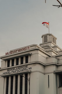 A historic building with art deco architecture features prominently, displaying the name 'Museum Mandiri' in bold red letters. Below is a sign with the words 'Nederlandsche Handel-Maatschappij N.V.'. An Indonesian flag flutters above the structure, which is characterized by clean lines and geometric shapes. The sky is overcast, adding an atmospheric quality to the scene.