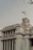 A historic building with art deco architecture features prominently, displaying the name 'Museum Mandiri' in bold red letters. Below is a sign with the words 'Nederlandsche Handel-Maatschappij N.V.'. An Indonesian flag flutters above the structure, which is characterized by clean lines and geometric shapes. The sky is overcast, adding an atmospheric quality to the scene.