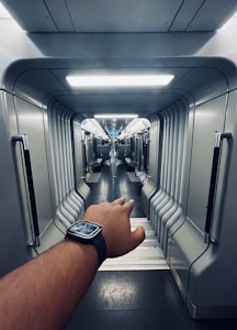 A futuristic train carriage interior with sleek, curved metallic lines and soft lighting. A person's hand wearing a smartwatch is reaching out into the aisle, suggesting motion or a journey.