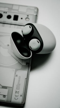 A pair of white wireless earbuds with black highlights is placed on top of a transparent-backed electronic device, possibly a smartphone or a tablet. The earbuds are encased in their charging case.