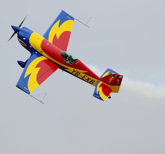 A brightly colored aerobatic airplane is captured in mid-flight against a clear sky, featuring a vibrant design with bold primary colors including blue, red, and yellow. Smoke trails can be seen extending from the aircraft during its aerial maneuver.
