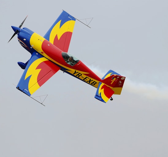 A brightly colored aerobatic airplane is captured in mid-flight against a clear sky, featuring a vibrant design with bold primary colors including blue, red, and yellow. Smoke trails can be seen extending from the aircraft during its aerial maneuver.