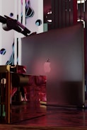 A sleek, modern setup featuring a laptop with an apple logo placed within an abstract, futuristic environment. Surrounding elements include glossy, reflective surfaces with a mix of red and metallic tones. Artistic, liquid-like forms and a telescope are present in the background, creating a surreal and technological composition.