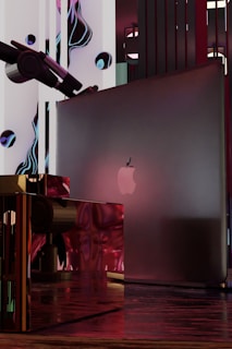 A sleek, modern setup featuring a laptop with an apple logo placed within an abstract, futuristic environment. Surrounding elements include glossy, reflective surfaces with a mix of red and metallic tones. Artistic, liquid-like forms and a telescope are present in the background, creating a surreal and technological composition.