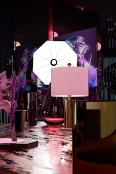 A modern interior space featuring a large illuminated octagonal light in the center, surrounded by abstract art pieces with vibrant colors. There's a contemporary lamp and reflective surfaces that create an artistic atmosphere.