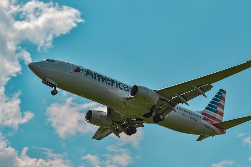 A passenger airplane with a visible logo soars through a vibrant blue sky scattered with fluffy white clouds. The aircraft's engines and landing gears are visible, indicating that it is either taking off or preparing to land.