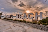Large sign spelling out 'Patong Beach' stands against the backdrop of a serene ocean view during sunset. The sky is filled with dramatic clouds as the sun casts a warm glow over the scene. Green plants line the base of the sign, and the area is paved with concrete.