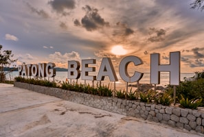 Large sign spelling out 'Patong Beach' stands against the backdrop of a serene ocean view during sunset. The sky is filled with dramatic clouds as the sun casts a warm glow over the scene. Green plants line the base of the sign, and the area is paved with concrete.