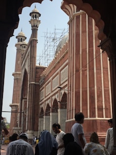 A historical mosque featuring tall minarets and arches. Scaffolding is visible, suggesting renovation work on the structure. Several people stand in the foreground, some wearing traditional garments, contributing to the cultural atmosphere.