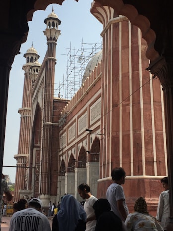 A historical mosque featuring tall minarets and arches. Scaffolding is visible, suggesting renovation work on the structure. Several people stand in the foreground, some wearing traditional garments, contributing to the cultural atmosphere.
