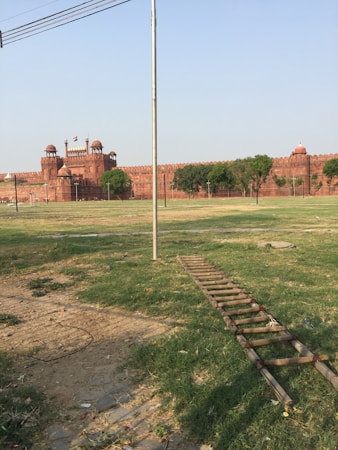 A large historic red sandstone fort is surrounded by green grass and trees. A flag flies atop the fort, which is adorned with domed structures. The foreground consists of a grassy field and a wooden ladder lying on the ground.