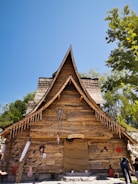 A rustic wooden house with a steep, pointed roof, adorned with Halloween decorations including hanging bats, a skeleton, and jack-o'-lanterns. The scene is set under a clear blue sky with green trees surrounding the building.
