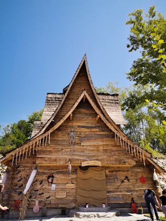 A rustic wooden house with a steep, pointed roof, adorned with Halloween decorations including hanging bats, a skeleton, and jack-o'-lanterns. The scene is set under a clear blue sky with green trees surrounding the building.
