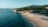 A serene beachfront with a stretch of sandy shore and calm blue waters. Pine trees cover the hillside in the background, while beach chairs and umbrellas are scattered across the sand. A small building with a sign reading 'OASIS' is nestled among the trees.