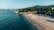 A serene beachfront with a stretch of sandy shore and calm blue waters. Pine trees cover the hillside in the background, while beach chairs and umbrellas are scattered across the sand. A small building with a sign reading 'OASIS' is nestled among the trees.