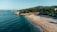 A serene beachfront with a stretch of sandy shore and calm blue waters. Pine trees cover the hillside in the background, while beach chairs and umbrellas are scattered across the sand. A small building with a sign reading 'OASIS' is nestled among the trees.
