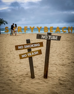 A sandy beach scene features large, colorful letters spelling 'SURFYBEACH' in the background near the ocean. In the foreground, there are wooden signs with messages prohibiting smoking, littering, and the use of tubes. A couple is visible walking along the beach under a cloudy sky, with a palm tree to the left.