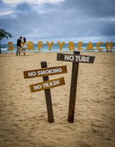 A sandy beach scene features large, colorful letters spelling 'SURFYBEACH' in the background near the ocean. In the foreground, there are wooden signs with messages prohibiting smoking, littering, and the use of tubes. A couple is visible walking along the beach under a cloudy sky, with a palm tree to the left.