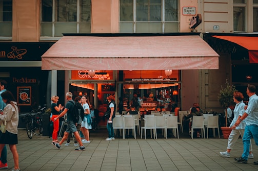 A lively street scene with people walking past an outdoor caf&eacute;. The caf&eacute; has an awning and serves as a gathering place for patrons sitting at white chairs and tables. The interior is warmly lit, creating a cozy atmosphere. Pedestrians are engaged in conversations or looking at their phones, adding a sense of everyday life.