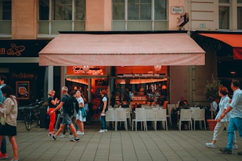 A lively street scene with people walking past an outdoor caf&eacute;. The caf&eacute; has an awning and serves as a gathering place for patrons sitting at white chairs and tables. The interior is warmly lit, creating a cozy atmosphere. Pedestrians are engaged in conversations or looking at their phones, adding a sense of everyday life.