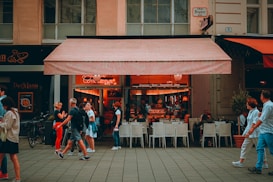 A lively street scene with people walking past an outdoor caf&eacute;. The caf&eacute; has an awning and serves as a gathering place for patrons sitting at white chairs and tables. The interior is warmly lit, creating a cozy atmosphere. Pedestrians are engaged in conversations or looking at their phones, adding a sense of everyday life.