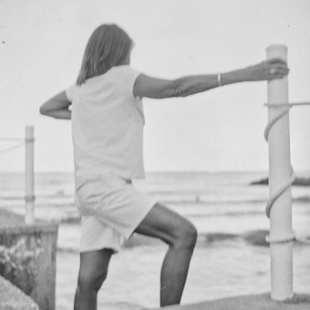 A person is stretching or exercising by holding onto a pole near the edge of a body of water. The black and white image creates a nostalgic and serene atmosphere. The person's pose and the surrounding scenery suggest a moment of calm and focus.