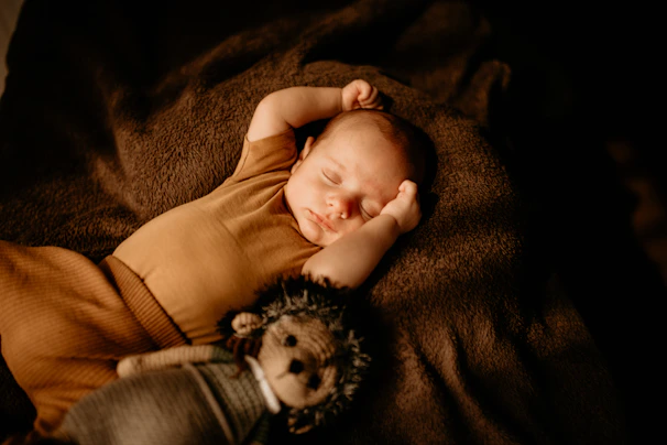 A peaceful sleeping baby lies on a soft, dark brown blanket. The infant is wearing a light brown outfit and appears relaxed with one arm raised near their face. A plush toy shaped like a hedgehog is placed next to the baby, adding a comforting presence.