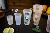 Four decorative cups with various patterns are arranged in a row on a dark surface. The designs include hearts, geometric shapes, a toucan with leaves, and polka dots. Around the cups, there are lime slices, mint leaves, small grey cubes, and partially visible fruits in the background.