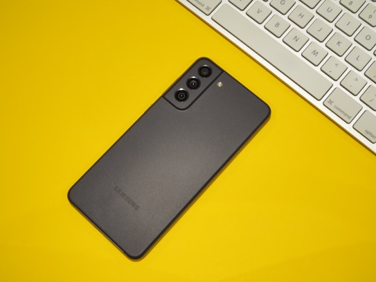 A black Samsung smartphone is placed on a bright yellow surface next to a white keyboard. The phone is positioned diagonally with its back facing up, displaying three camera lenses and a flash.