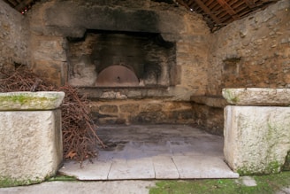 A rustic stone oven is built into a rugged stone structure with a curved dome. The surrounding walls and roof are made of weathered stone and wood, suggesting an old, possibly abandoned building. Dried twigs or small branches are piled up in front of the oven, likely for fuel.